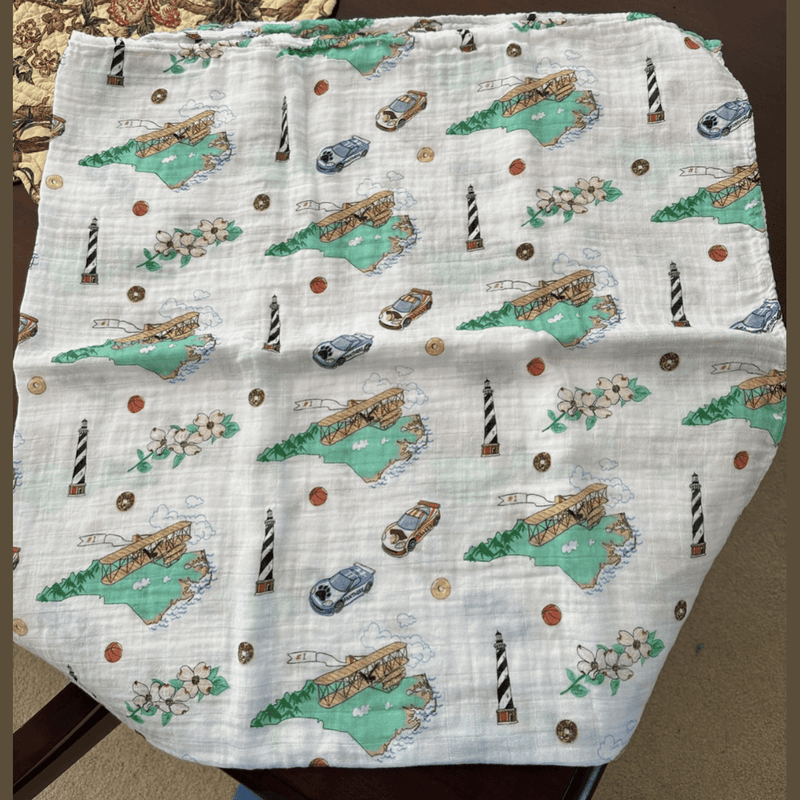 North Carolina-themed baby gift set with muslin swaddle blanket, burp cloth, and bib in soft pastel colors.