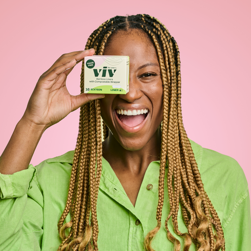 Gen-Z woman in a green shirt holding a box of panty liners in front of her eye, she is smiling and looking content.