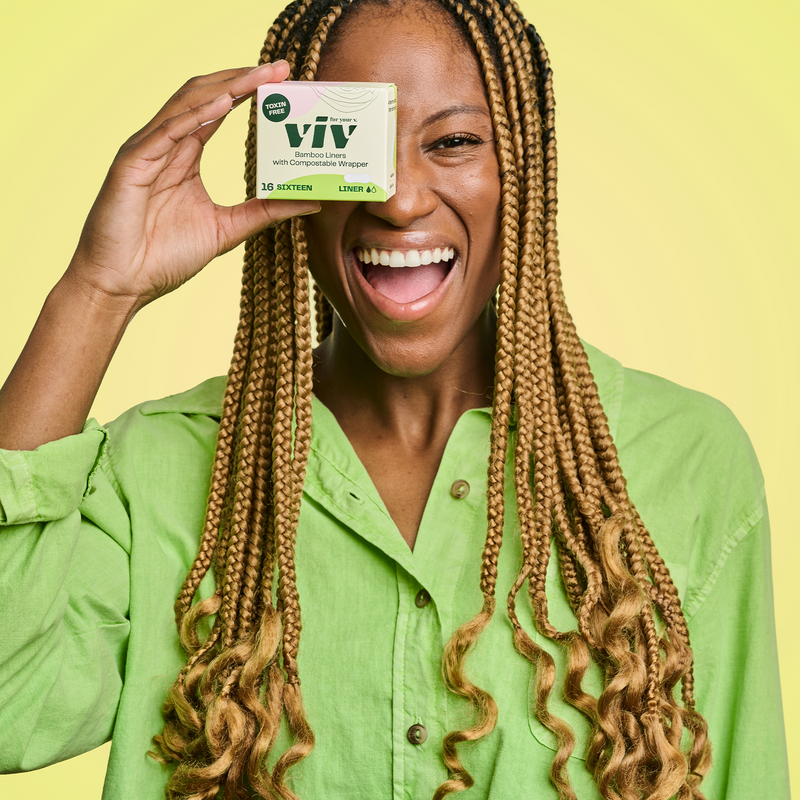 Gen-Z woman in a green shirt holding a box of panty liners in front of her eye, she is smiling and looking content.