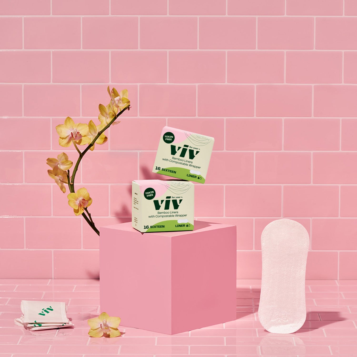 A set of two boxes with biodegradable bamboo liners resting on a pink box in a pink tile bathroom with flowers in the background. A sustainable panty liner is seen standing up. 