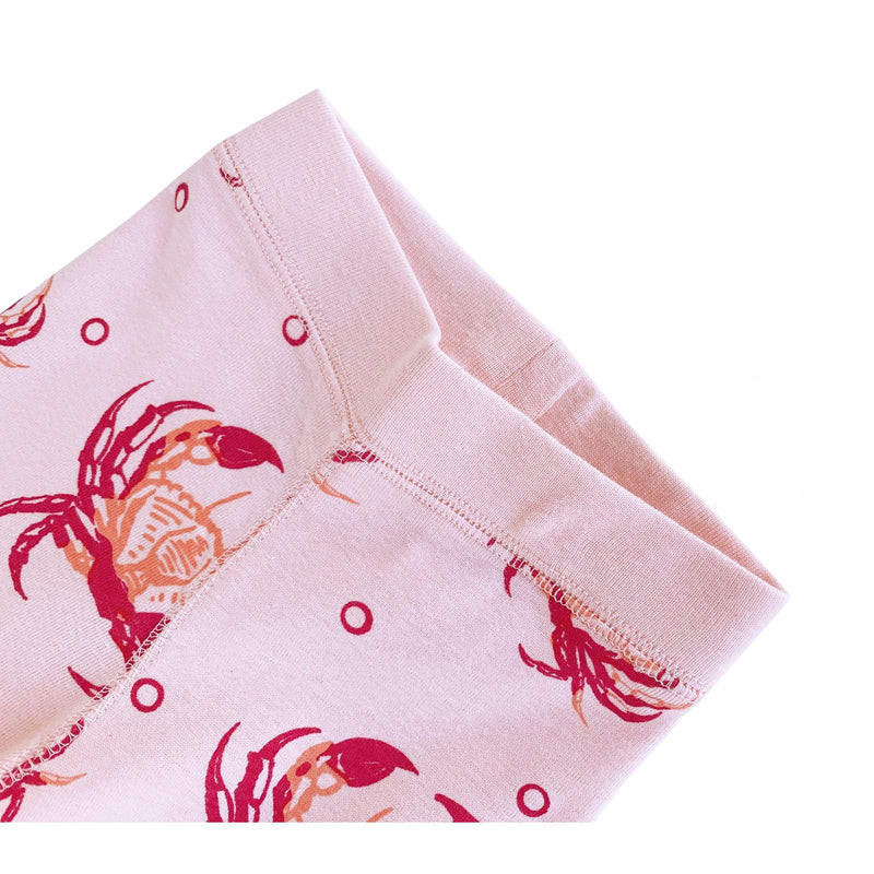 Toddler wearing pink pajamas with cute crab patterns, smiling and playing on a white bed with a plush toy.